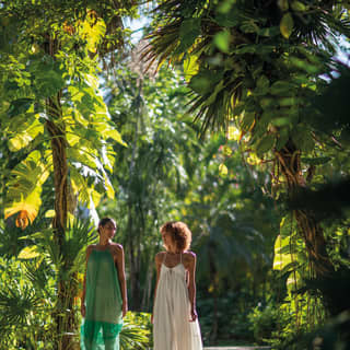 Two female guests in green and white diaphanous dresses walk through tree ferns and tropical plants in the jungle gardens.