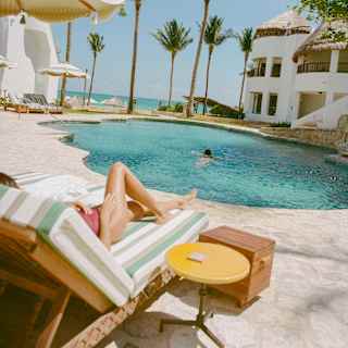 A lone swimmer enjoys the rippling blue tropical pool, seen over a lounger where a female sun-bather's legs stretch out.