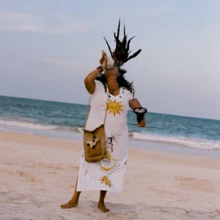 A shaman, in a white dress with a sun emblem and black feather headdress, blows a conch horn at a ceremony on the beach.