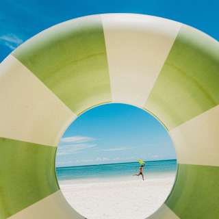 A person is jumping on a sandy beach, seen through the centre of a large green and white striped inflatable swimming ring with the sea and blue sky in the background.