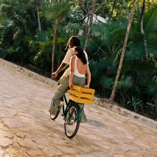 A woman rides on the back of a bike, holding onto a yellow box behind her as the male rider sets off along the stone path.