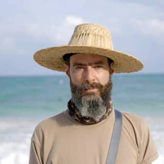 Portrait of photographer Ilán Rabchinskey, with dark hair and greying beard, wearing a raffia hat with his back to the sea.