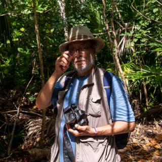 Photographer Javier Hinojosa, with short grey beard, glasses and sun hat stands in a jungle setting holding his camera.