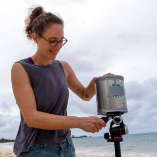 Photographer Margot Kalach, with her hair tied up, wearing a blue vest top and glasses, sets up her equipment on the beach.