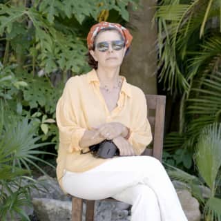 Photographer Patricia Lagarde, seated outside with a camera in her lap, wears a red head scarf, sunglasses and yellow shirt.