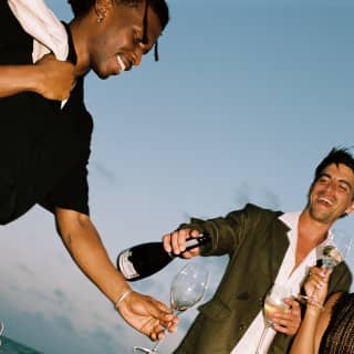 A male guests pours wine for a friend extending his glass as they enjoy a celebration on Pescados Beach, shot at an angle.