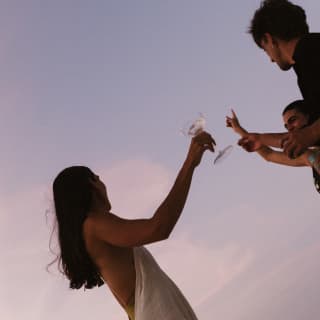Angled shot of a long-haired woman in a white dress as she raises a glass with two male friends, taken against a lilac sky.