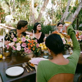 Seen from behind, a woman in a green top raises a toast with friends at flowery celebration table in the leafy courtyard.