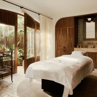 A white bed bathes in light from the large windows of a spa room with wood panels, towel rack, marble sink and garden views.