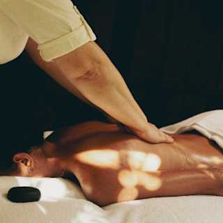 A therapists performs a massage on the oiled back of a guest, who lies on a comfortable treatment bed in dappled sun.