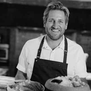 Chef Curtis Stone smiles to camera, wearing a dark apron and white shirt, holding plates of seafood in a monochrome image.