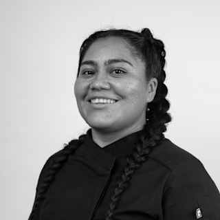 Oaxacan chef, Thalía Barrios García, smiles to camera in a dark top with her long black hair in two plaits, in monochrome.