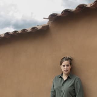 In a green shirt, with hair tied up, Mauka chef Pía León stands against a rendered ochre wall with terracotta-tile roof.