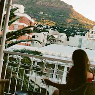 A woman sits with a drink on the monochrome chequered Strelitzia Suite balcony, gazing at the mountains, seen from behind.
