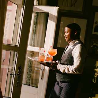 Sun through open doors catches the peachy glow of four cocktails on a tray carried by a waiter in a black waistcoat.