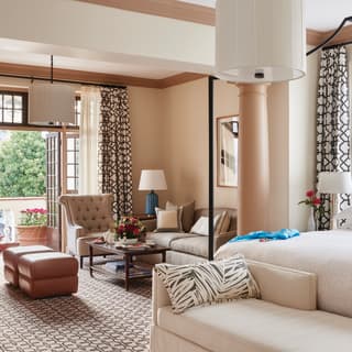 View over a Junior Suite bed to a retro-chic living space with beige sofa and chair, swirly carpet and caramel-cream walls.