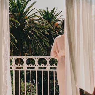 A woman in a pink robe leans on the white railings of her balcony, gazing over lush palm trees, seen from behind a curtain.