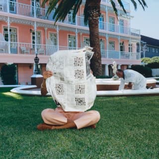 On the lawn outside tiers of suites with pretty white-rail balconies, a woman reads a large newspaper that covers her face.