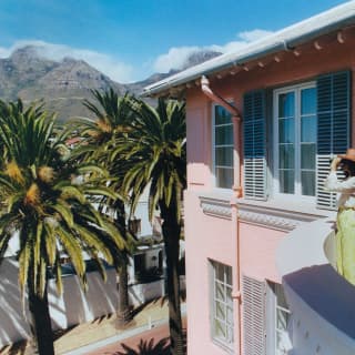 A woman in a brown hat, white shirt and green trousers stands on her suite balcony looking at the view through binoculars.