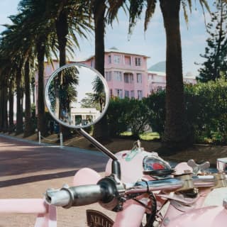 A vintage, pink side-car motorcycle points towards the hotel, with the gateway entrance in the rear view mirror.