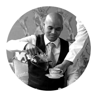 Black and white porthole photo of a male staff member, in black waistcoat and tie, pouring tea into a cup, held by a waiter.