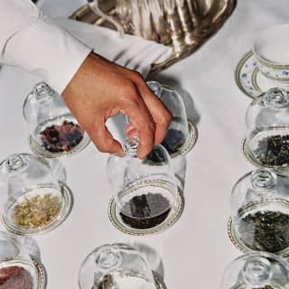 The hand of a tea sommelier selects one of nine varieties of tea leaves, displayed on small plates beneath glass cloches.