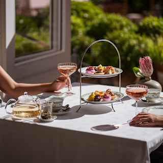 Two guests, out of shot, enjoy glasses of rosé at an Afternoon Tea table with a two-tier patisserie stand, in bright sunshine.
