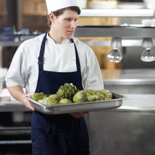 Kitchen scene with a young chef, wearing a navy apron and a white toque, carrying a metal tray of vibrant green artichokes.