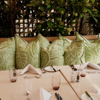 View over a dining table with with beige-cream cloth and bench seating with botanical print green cushions at Oasis.