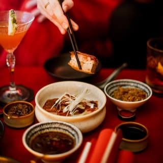 A woman lifts a piece of duck with black chopsticks at a table with a cocktail and dipping bowls at Red Room restaurant.