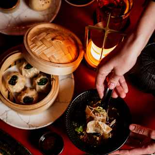 A guest reaches for a bowl of gyozas at a Red Room table busy with Asian dishes like dumplings and bao buns, seen from above.