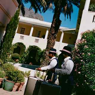 Angled image of two male members of staff, in uniform and hats with pink ribbons, as they wheel suitcases towards the hotel.