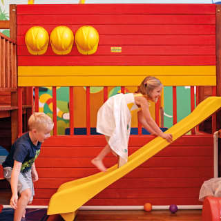 Two kids climbing a yellow slide in a brightly coloured kid's playroom