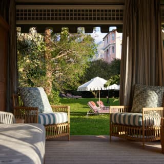 Two pink loungers recline under a white parasol on the lawn, seen from a cabana with open drapes and three lounge chairs.