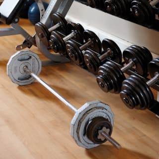 Fitness centre detail of a weights rack with rows of black dumbbells and a long silver weighted barbell on the floor.