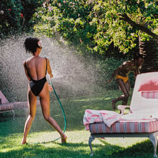 A woman in a black swimming costume sprays a male companion with a hose as he runs in yellow trunks beneath a garden tree.