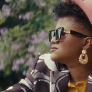 Fashion designer Palesa Mokubung wears a pink hat and dark glasses with a yellow shirt and bold brown and white print jacket.