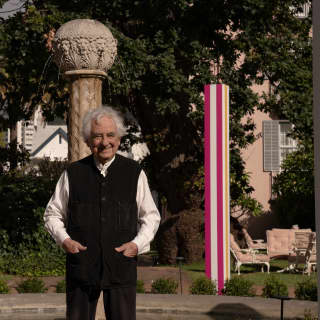 Celebrated artist Daniel Buren stands between pink and white obelisks in a black waistcoat, smiling with hands in pockets.