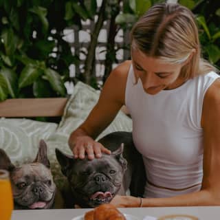 A blonde woman in a white top pats two French Bulldogs on the bench next her on, seen over a table with coffee and a pastry.