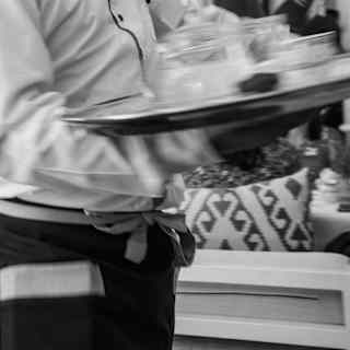A waiter carries a tray, blurred by motion, as he serves during a party, seen close-up in black and white.