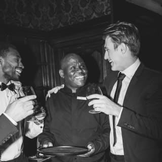 Black and white image of a party as a suited man with a drink rests one arm on the shoulder of a waiter as they share a joke.