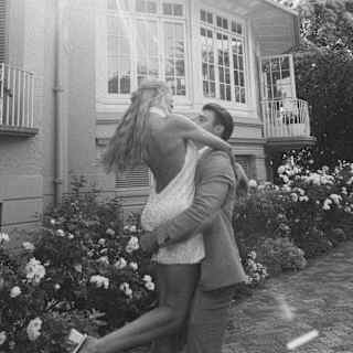 In a flowering garden, a suited man lifts a woman with long blonde hair and a low-back white dress, captured in monochrome.