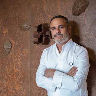 Amura Head Chef Ángel León, wearing chef whites, stands against a copper-hued wall, smiling to camera with arms folded.