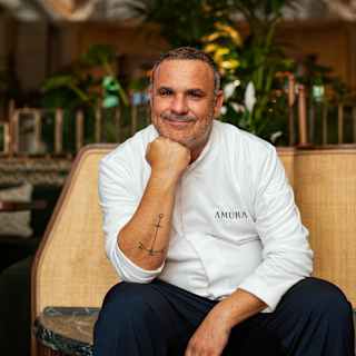 A smiling chef in a white uniform sits on a beige bench with his hand resting on his chin, indoors with plants and warm lighting in the background.