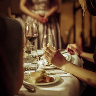 A woman rests her wrists on the edge of a dining table, holding a piece of bread as she turns to watch the entertainment.