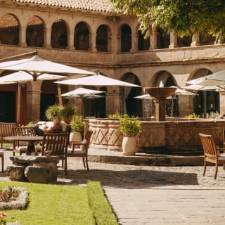 Diagonal view over the courtyard where white umbrellas and outdoor tables circle a fountain, with the cloisters behind.