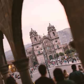 Angled low-light shot of crowds outside Iglesia de la Compañia de Jesús in Cusco's Plaza Mayor, seen through arcade arches.
