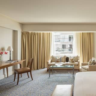 A cream sofa and chair enjoy city views by a window with open wheat-tone drapes in a Deluxe Junior Suite with a desk and bed.