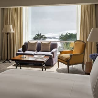 A brown sofa and coffee table bathe in light from a window over the ocean, seen from the bed of an Ocean View Junior Suite.