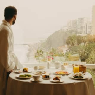 Low-saturation image of a man in a bathrobe on the balcony of his suite by a table with breakfast and a breathtaking view.